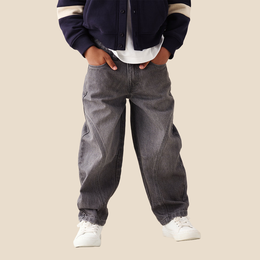 A child wearing baggy dark gray washed denim jeans with contrast stitching, styled with a navy and beige varsity jacket, white t-shirt, and white sneakers against a solid beige background.