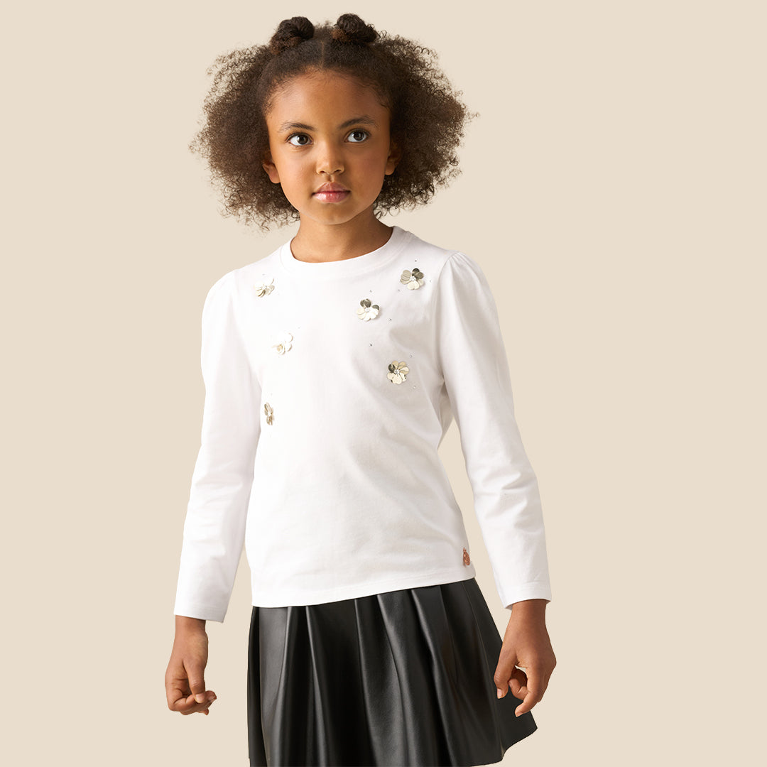 A young girl with curly hair wearing a white long-sleeve top with decorative metallic appliques, paired with a black skirt, against a solid beige background.