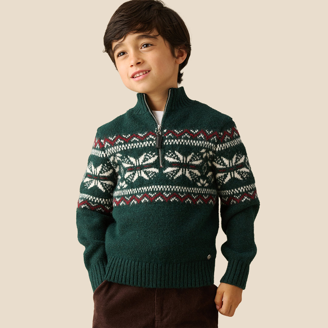 A young boy wearing a dark green knit half-zip sweater with a white and burgundy Fair Isle pattern across the chest and sleeves, paired with dark brown pants, set against a solid beige background.