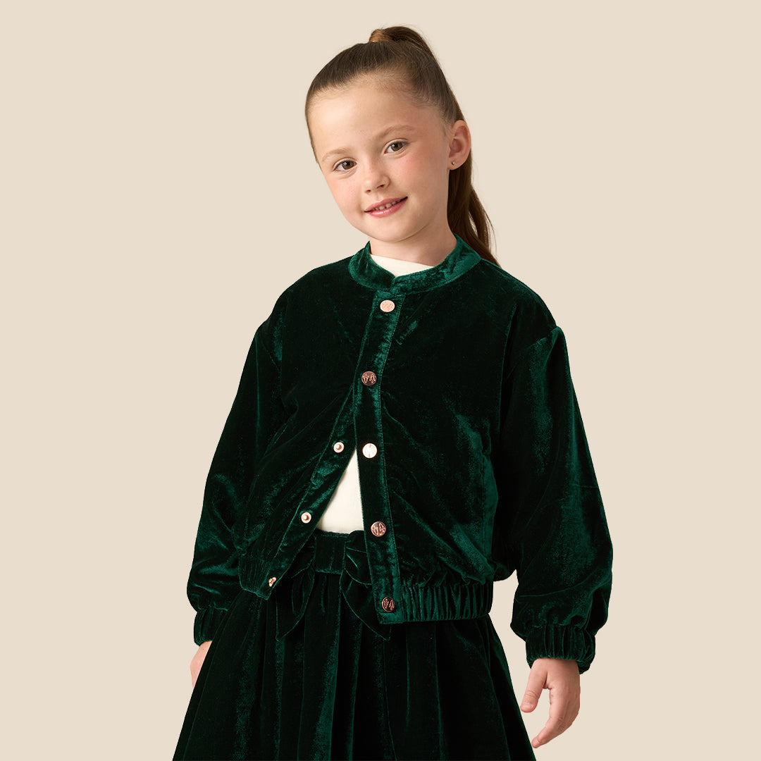 A young girl with her hair in a ponytail wearing a dark green velvet jacket and matching skirt over a white top, photographed against a solid beige studio background.