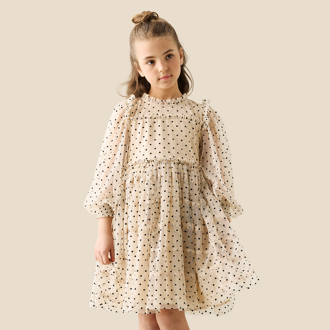 A young girl with her hair in a bun wears a cream-colored, long-sleeved tiered dress with black polka dots, standing against a solid beige background.