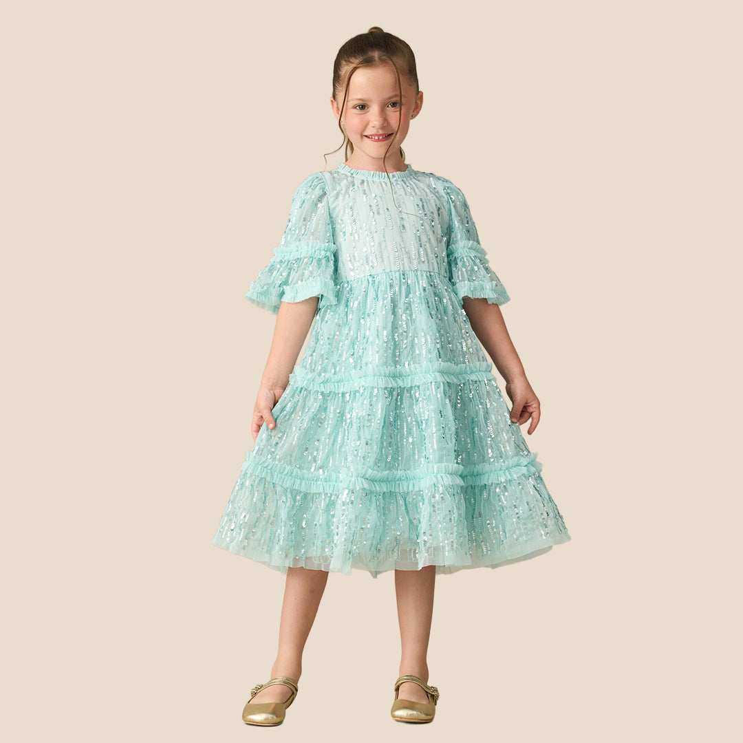 A young girl with a long brown ponytail wearing a light mint green sequined tiered dress, standing against a solid beige background.