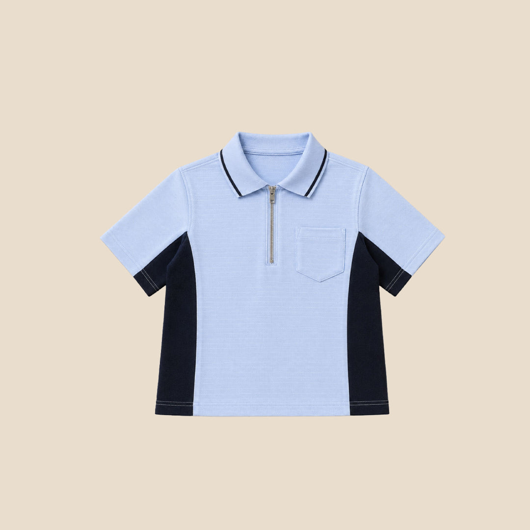 A young boy modeling a light blue polo shirt with dark navy side panels and a half-zip collar, paired with navy pants, against a solid beige background.