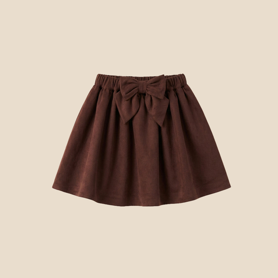 A model wearing a beige ribbed cardigan and a dark brown pleated skirt with a large bow at the waist, shown from the chest down against a solid beige background.