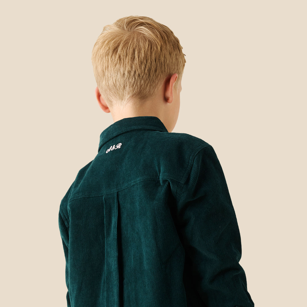 A young boy with blonde hair wearing a dark forest green corduroy button-up overshirt over a white t-shirt and matching green pants, standing against a solid beige background.