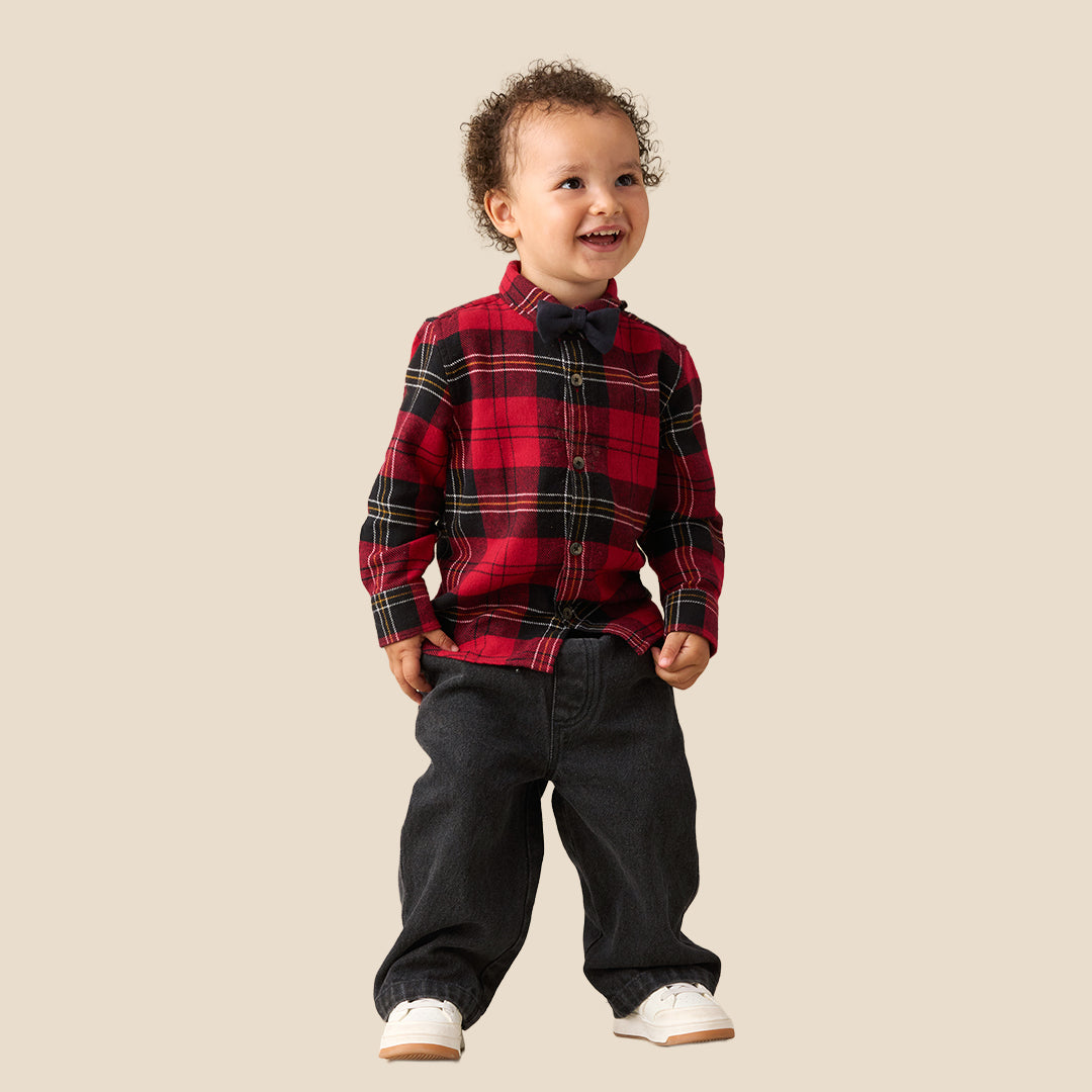 A toddler standing against a plain beige background, wearing a red and black plaid long-sleeve shirt with a matching dark bow tie, dark pants, and white sneakers.