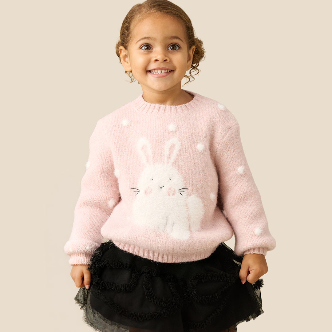 A young girl smiling while wearing a light pink knit sweater with a white bunny applique and white pom-pom details, paired with a black ruffled tutu skirt, against a solid beige background.