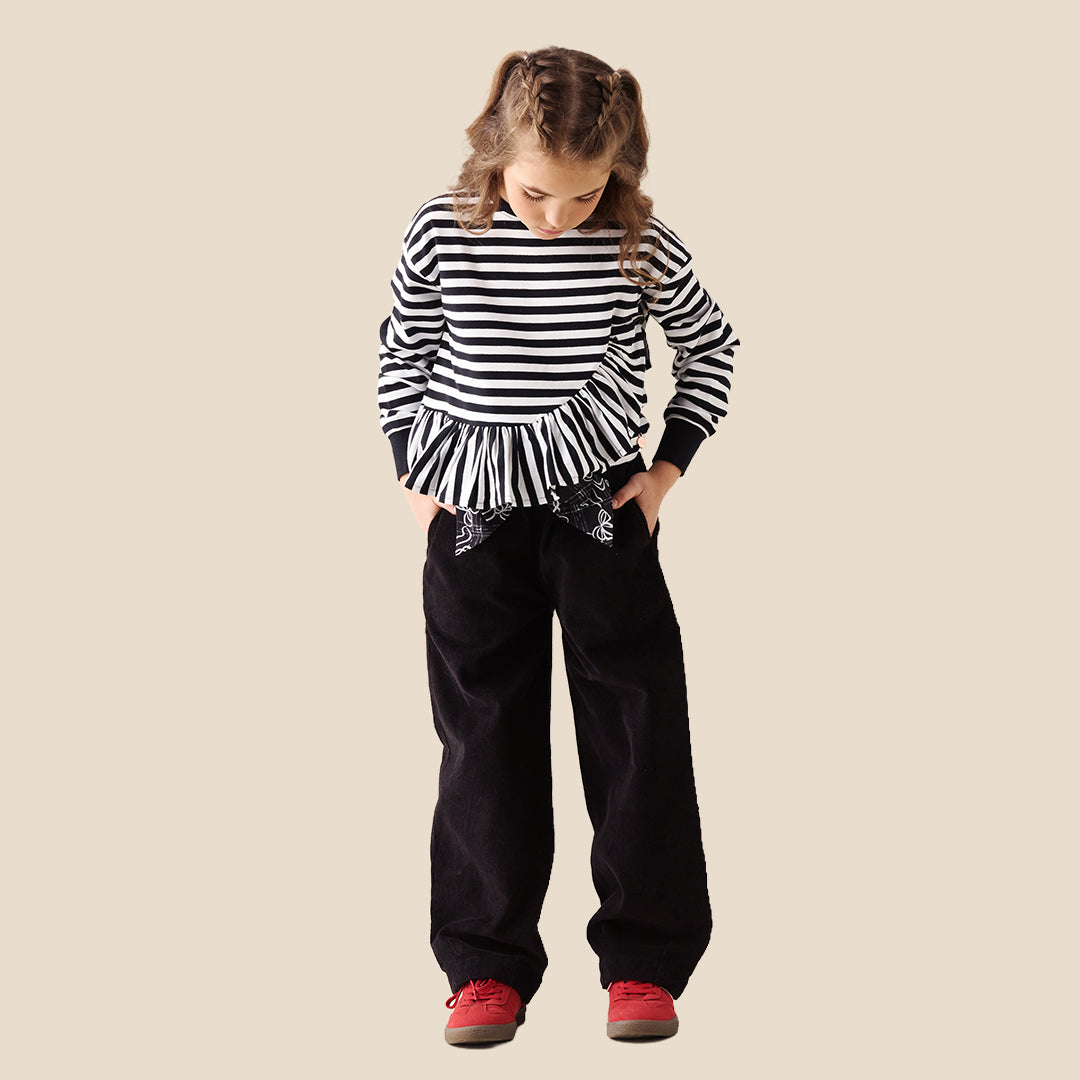 A child wearing black wide-leg pants with a white bow and floral pattern at the waistband, paired with a black top and red sneakers, standing against a solid beige background.