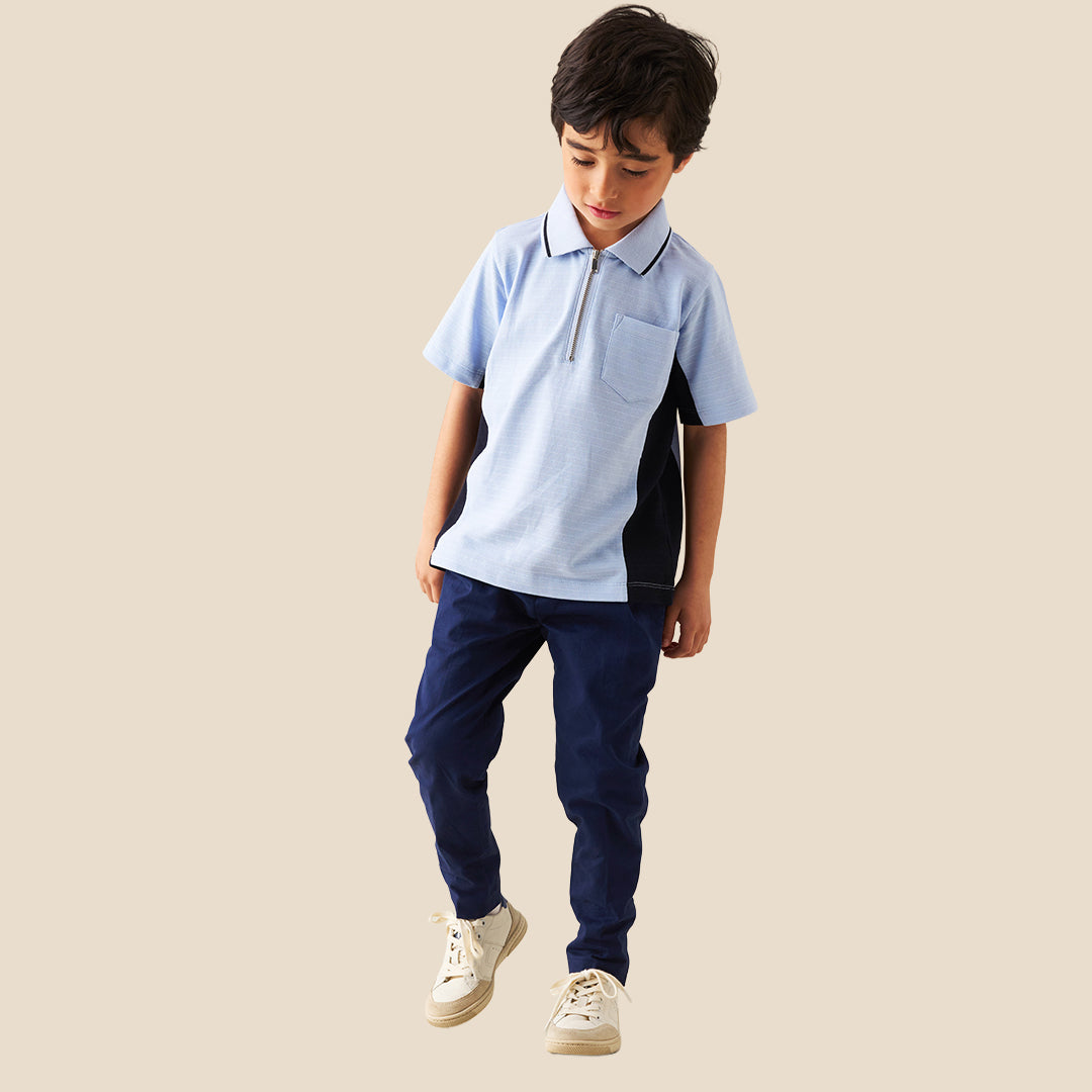 A young boy modeling a light blue polo shirt with dark navy side panels and a half-zip collar, paired with navy pants, against a solid beige background.