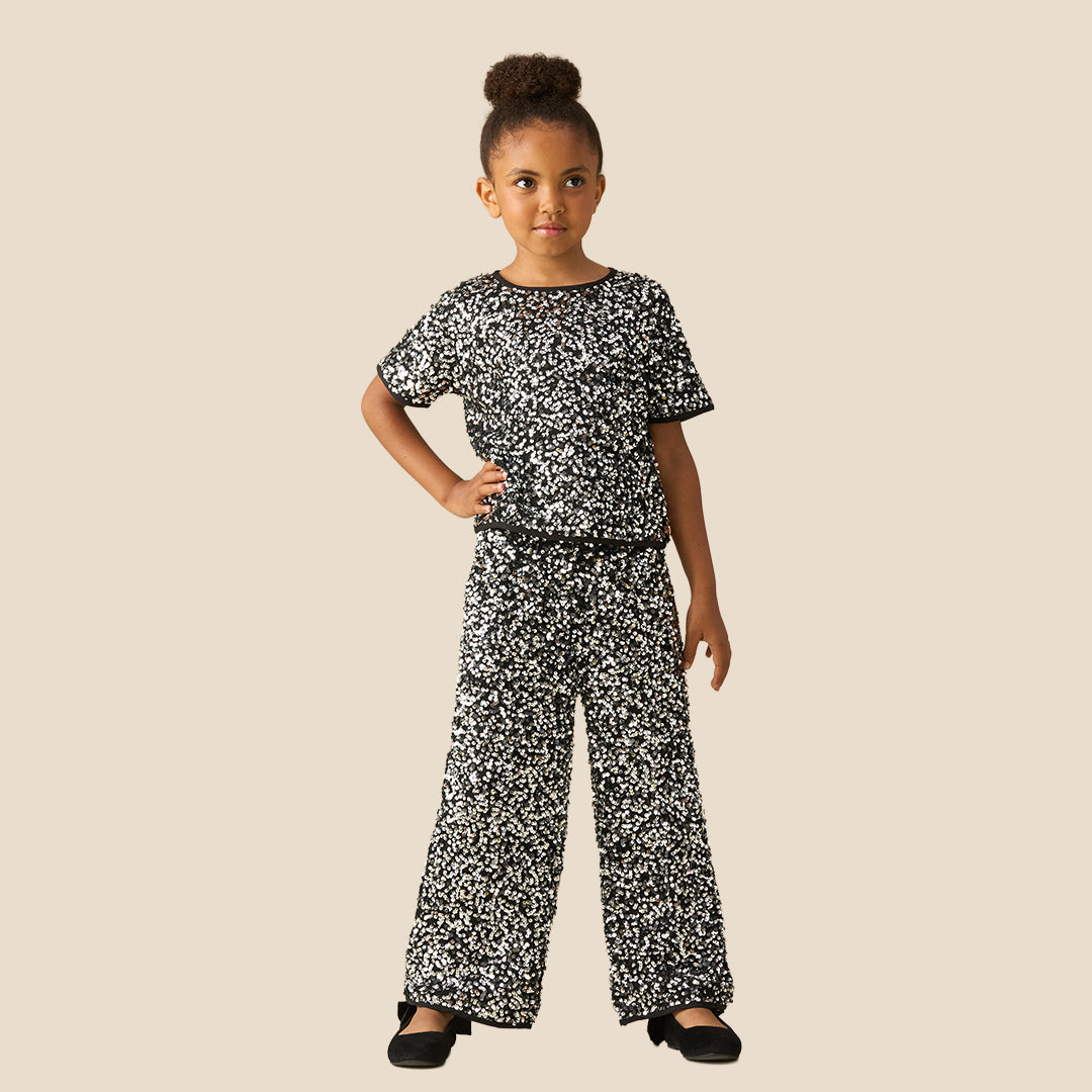 A child modeling a matching two-piece outfit with a black and white speckled pattern, featuring a short-sleeve top and wide-leg pants, styled with black flat shoes against a solid beige background.