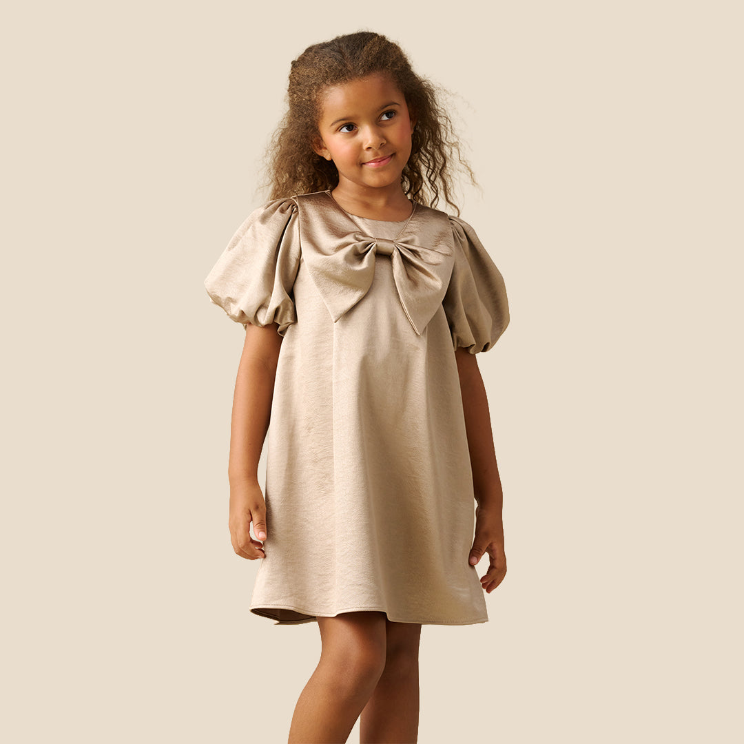 A young girl with curly brown hair wearing a champagne beige dress with puffed short sleeves and a large bow at the neckline, standing against a solid light beige background.