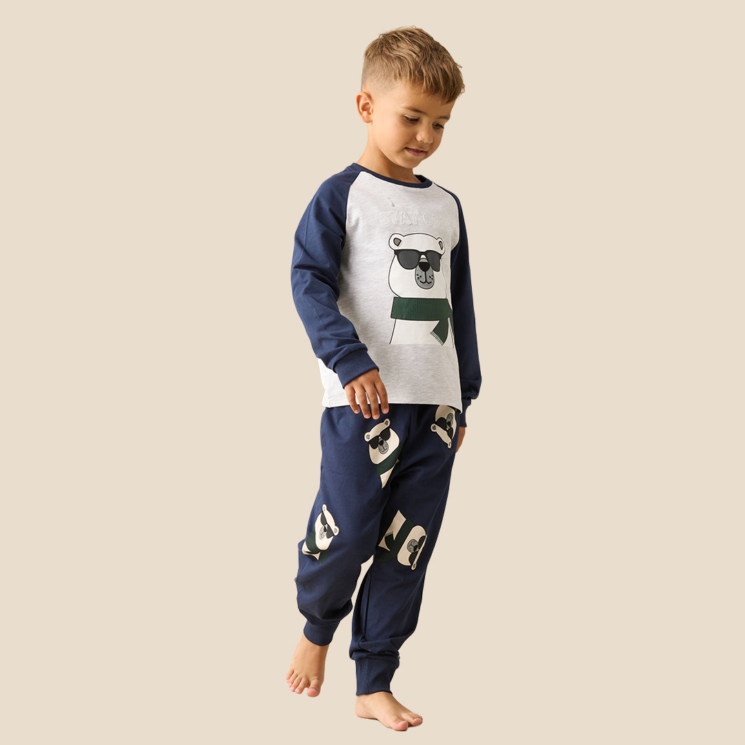 A young boy modeling a two-piece pajama set against a solid beige background. The set features a white long-sleeve top with navy blue raglan sleeves and a cartoon bear graphic wearing sunglasses and a green scarf, paired with navy blue pants decorated with patchwork-style graphic details.