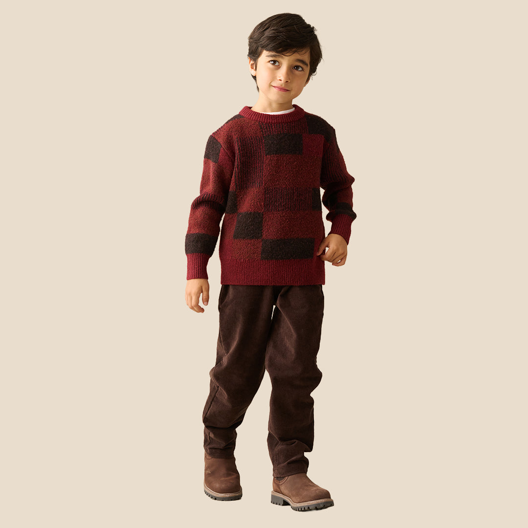 A young boy wearing a textured knit sweater with a dark red and black checkered pattern, paired with dark brown pants, set against a solid light beige background.
