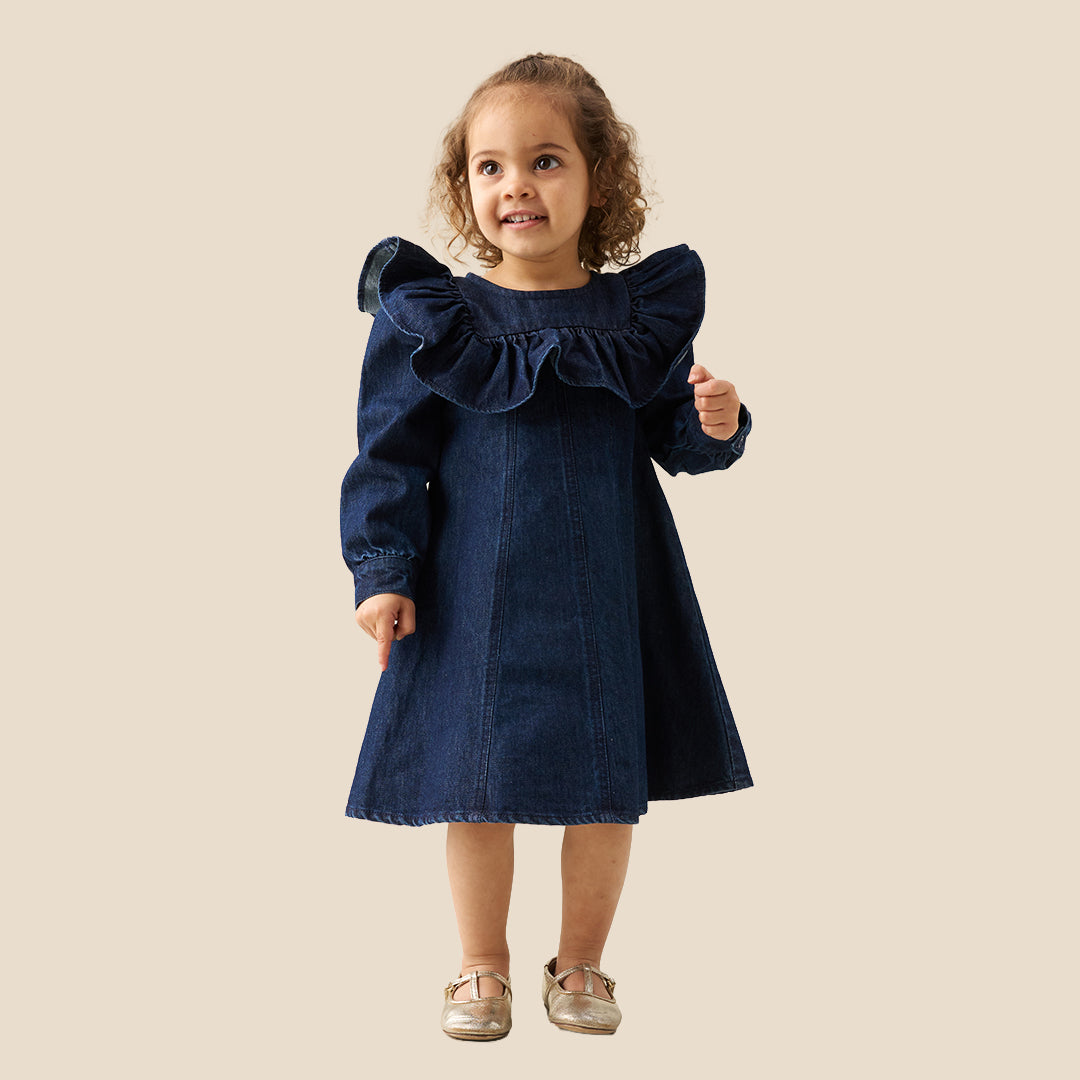 A young child wearing a dark blue denim dress with dramatic ruffled shoulders, standing against a solid beige background.