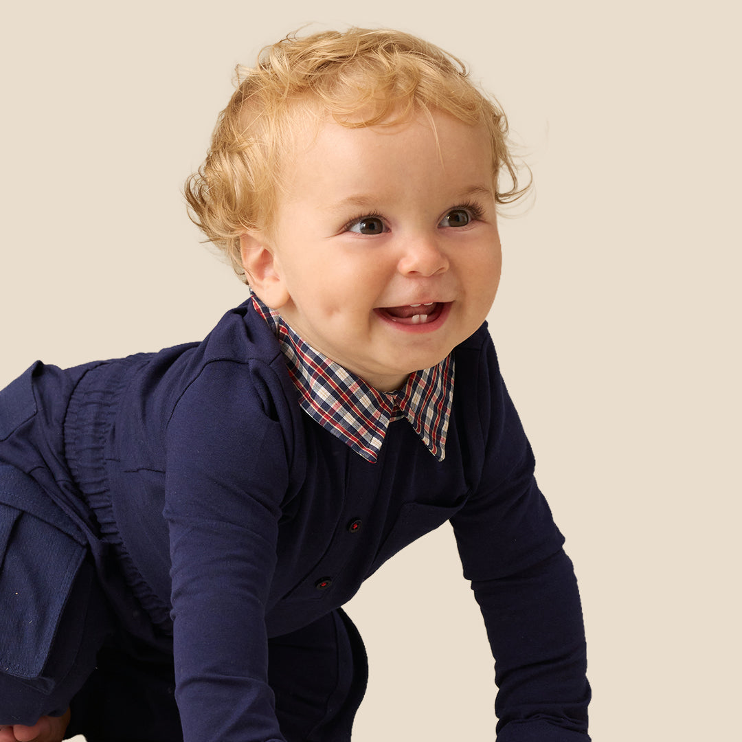A smiling toddler with curly blond hair wearing a navy long-sleeve shirt and matching pants over a red, white, and blue plaid collared shirt, photographed against a plain warm beige studio background.