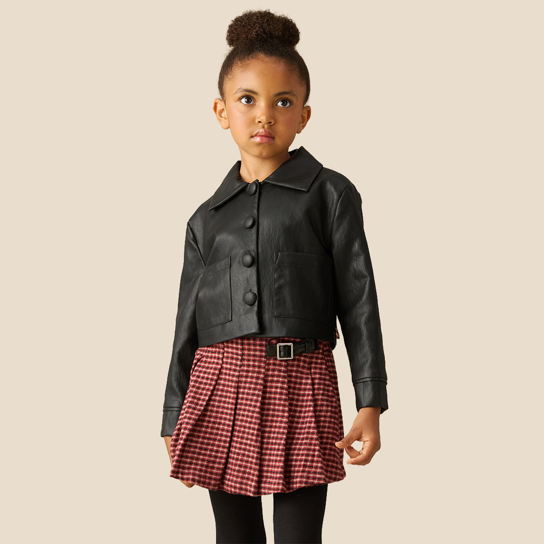 A close-up of a pleated skirt in a red and black gingham pattern, styled with a black leather jacket and black tights against a solid beige background.