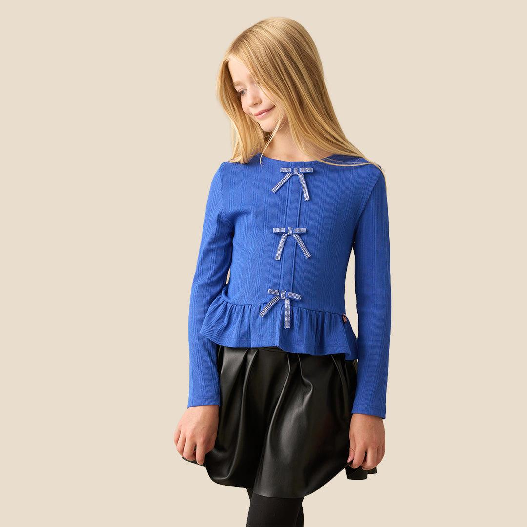 A young girl with long blonde hair wearing a blue ribbed long-sleeve top with three light blue bow details on the front and a black skirt, standing against a solid beige background.