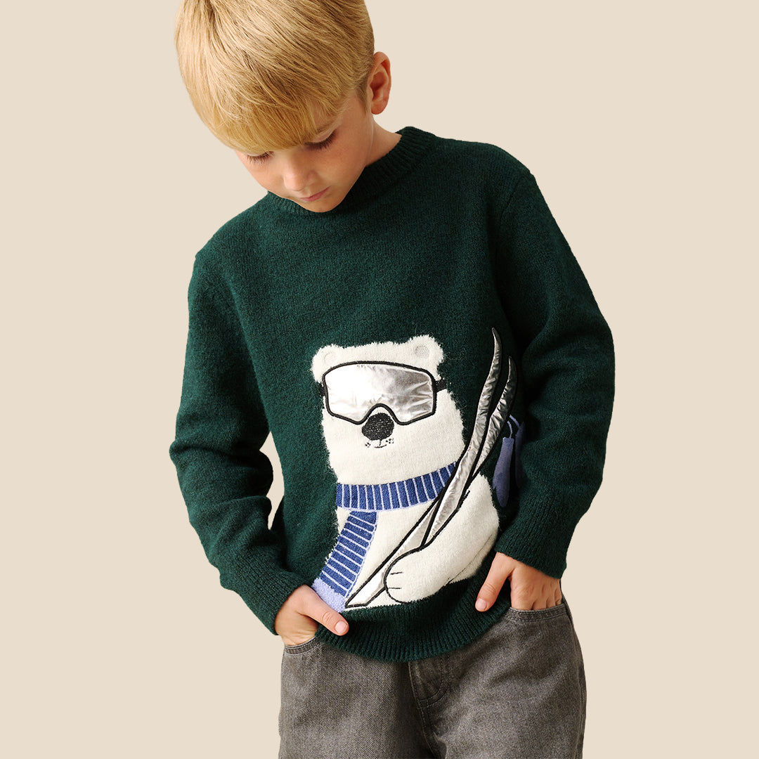 A young boy with blond hair wearing a dark green knit sweater featuring a large white polar bear graphic with ski goggles and a blue striped scarf, set against a solid beige background.