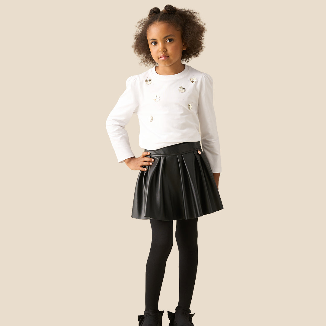 A child model wearing a black faux-leather pleated mini skirt with a matching black belt and rose gold button, paired with a white embellished top and black tights, against a solid beige background.