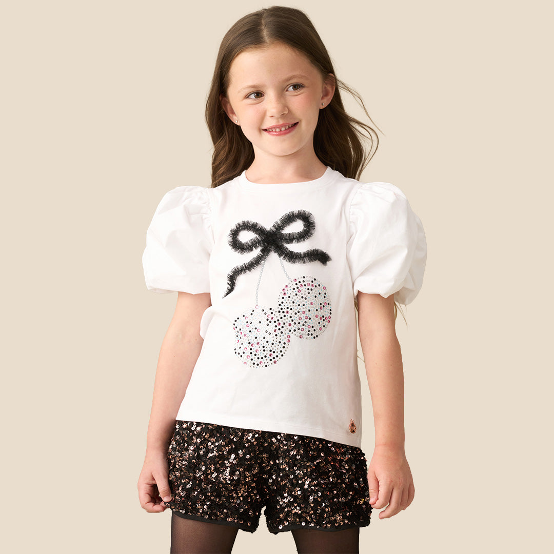 A young girl modeling a white short-sleeve top with puff sleeves and a sequined cherry graphic, paired with black sequined shorts, against a solid beige background.