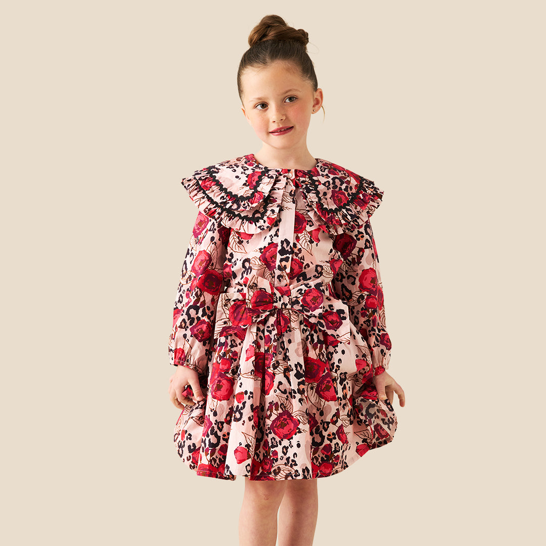 A young girl with her hair in a bun stands wearing a long-sleeve dress with a red rose and black leopard print pattern on a light pink background, set against a solid beige studio backdrop.
