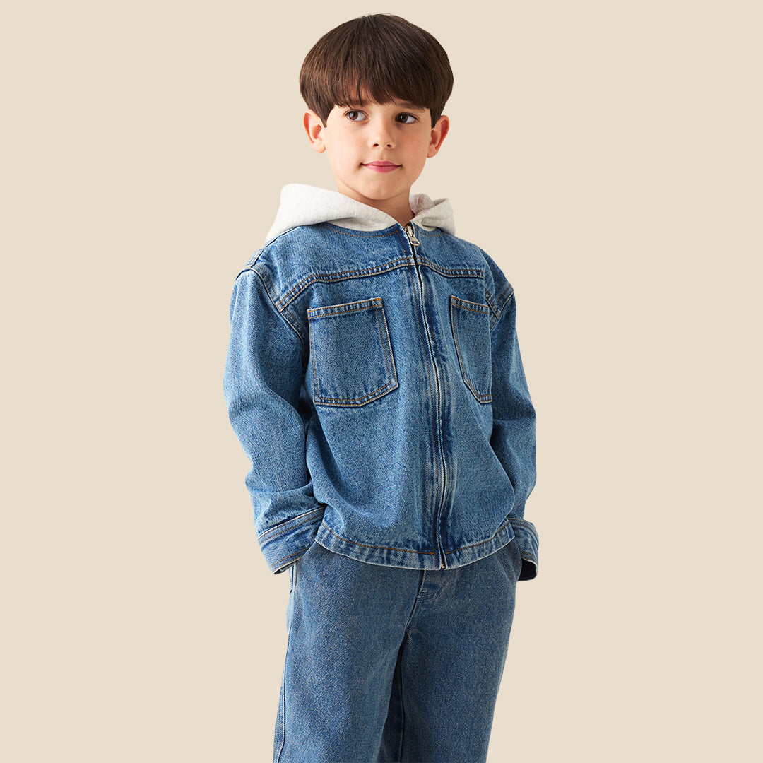 A young boy wearing a light blue denim zip-up jacket with a white hood lining and matching blue jeans, standing against a solid beige background.