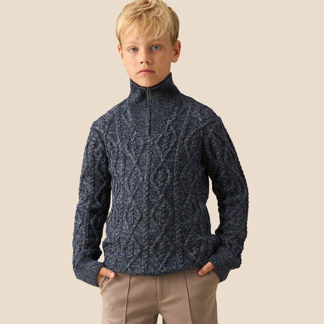 A young boy with blonde hair wearing a navy blue cable-knit mock-neck sweater and beige trousers, standing against a solid beige background.