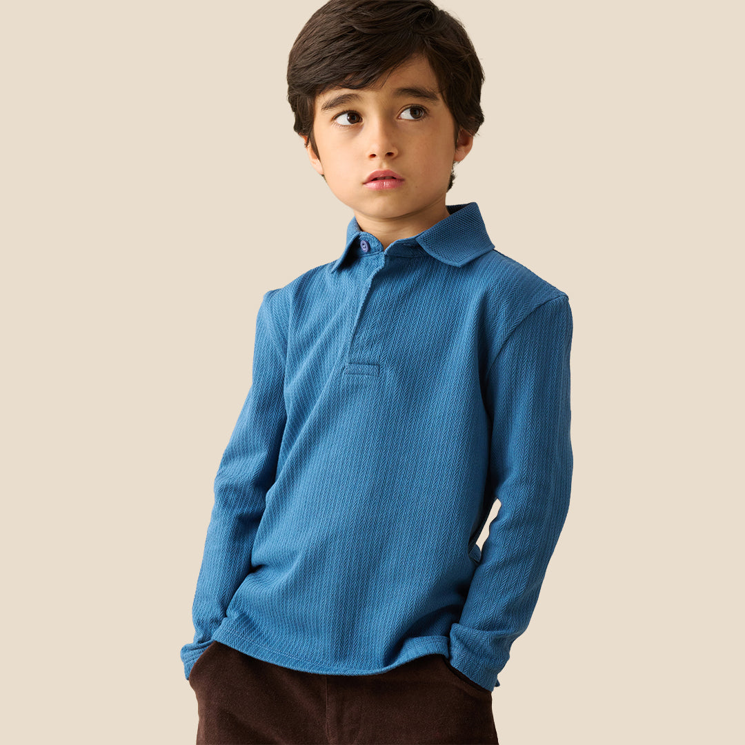 A young boy wearing a blue ribbed long-sleeve polo shirt and brown pants, standing against a solid beige background.