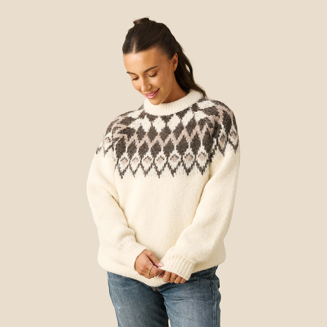 A woman wearing a cream-colored knit sweater with a dark gray and brown Fair Isle yoke pattern, paired with blue jeans, against a solid beige background.