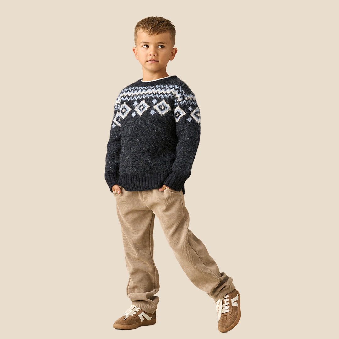 A young boy wearing a dark navy knit sweater with a white and light blue geometric Fair Isle pattern across the chest and shoulders, paired with beige pants, set against a solid light beige background.
