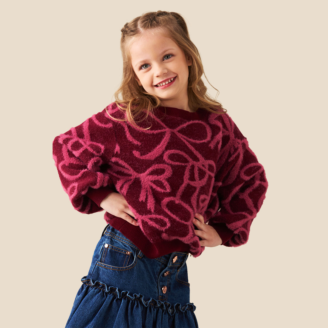 A young girl with blonde hair in a ponytail wearing a fuzzy dark red long-sleeve sweater with a pink bow pattern, paired with a blue denim skirt, against a solid beige background.