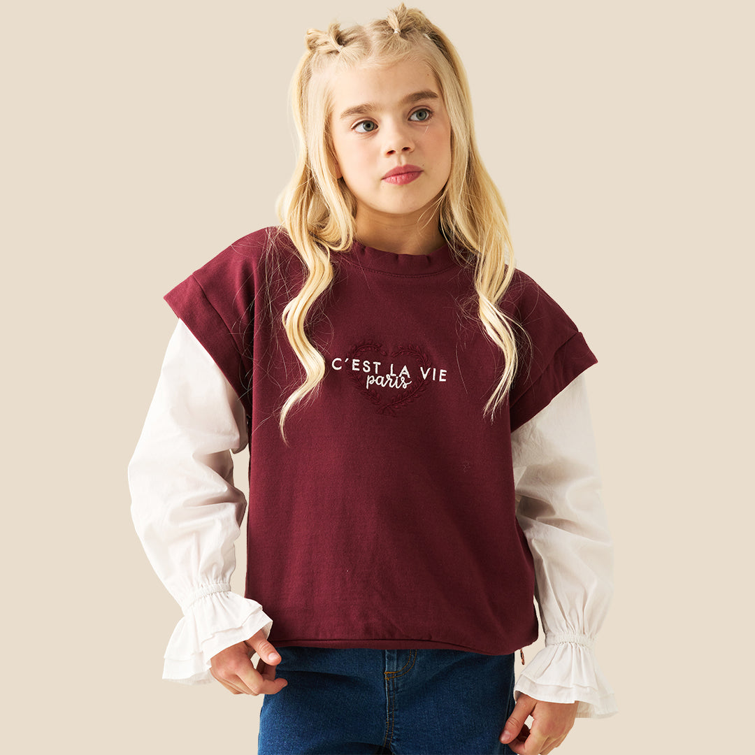 A young girl with blonde pigtails wearing a maroon cropped t-shirt with white sleeves and 'C'EST LA VIE paris' printed in white, layered over a white long-sleeve blouse with ruffled cuffs, and blue jeans, against a solid beige background.