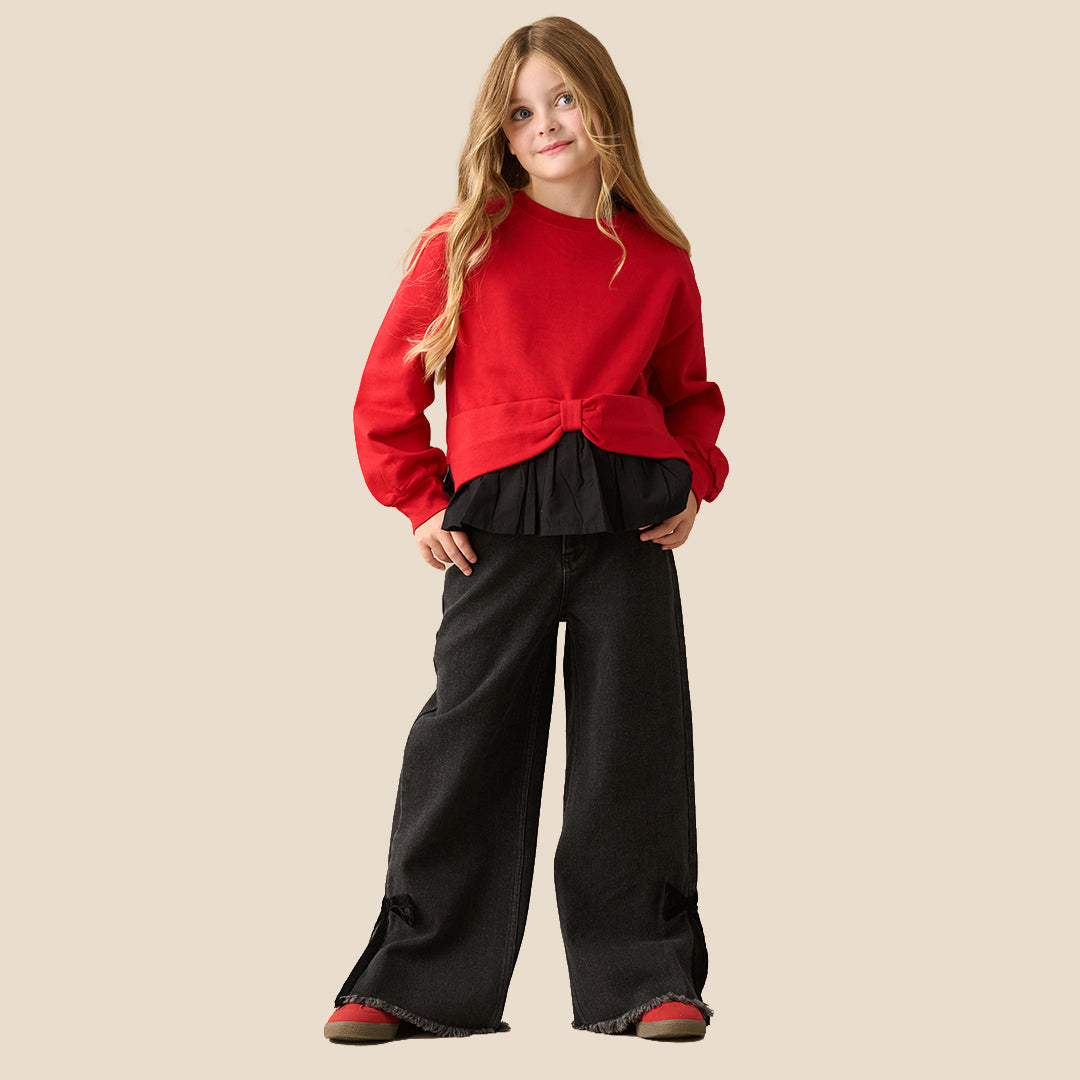 A person wearing black wide-leg denim jeans with side tie details and raw frayed hems, styled with a red bow-tie sweatshirt and red sneakers, against a solid beige background.