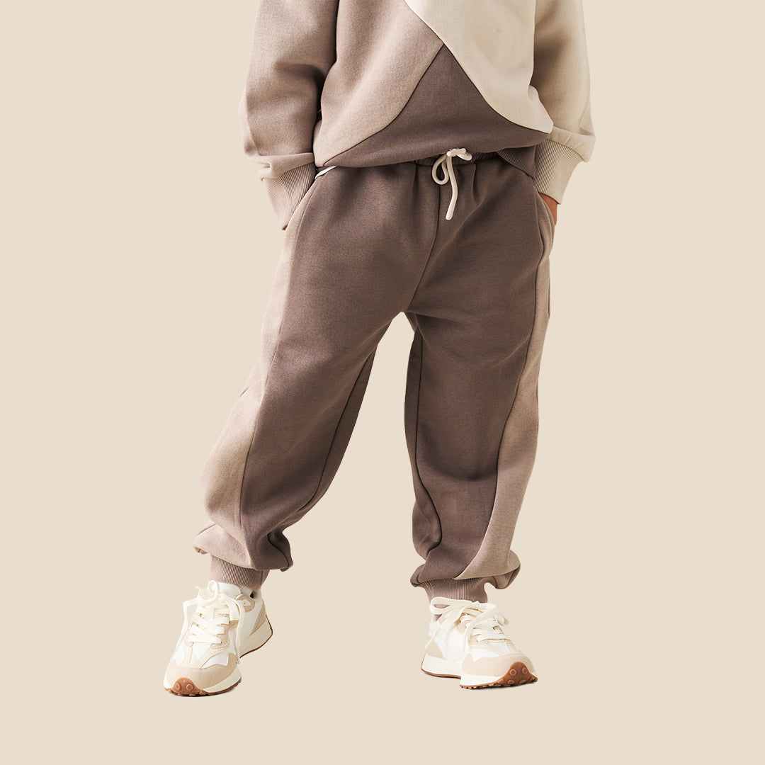 A person wearing loose taupe-brown jogger sweatpants with a white drawstring, styled with a beige and cream sweatshirt and white sneakers, against a solid beige background.
