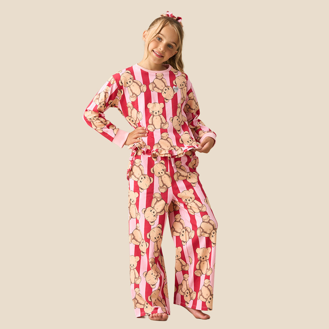 A young girl modeling a two-piece pajama set featuring a pink and white striped pattern with a repeating teddy bear print, set against a solid beige background.