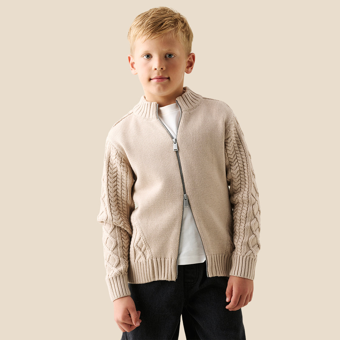 A young boy wearing a beige cable-knit zip-up cardigan over a white t-shirt and dark jeans, standing against a solid light beige background.