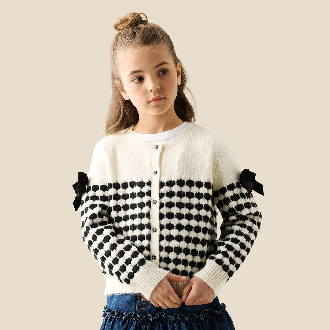 A young girl with her hair in a bun wears a cream knit cardigan with black polka-dot patterned sleeves and black ribbon bows on the shoulders, paired with a dark blue ruffled skirt, against a solid beige background.