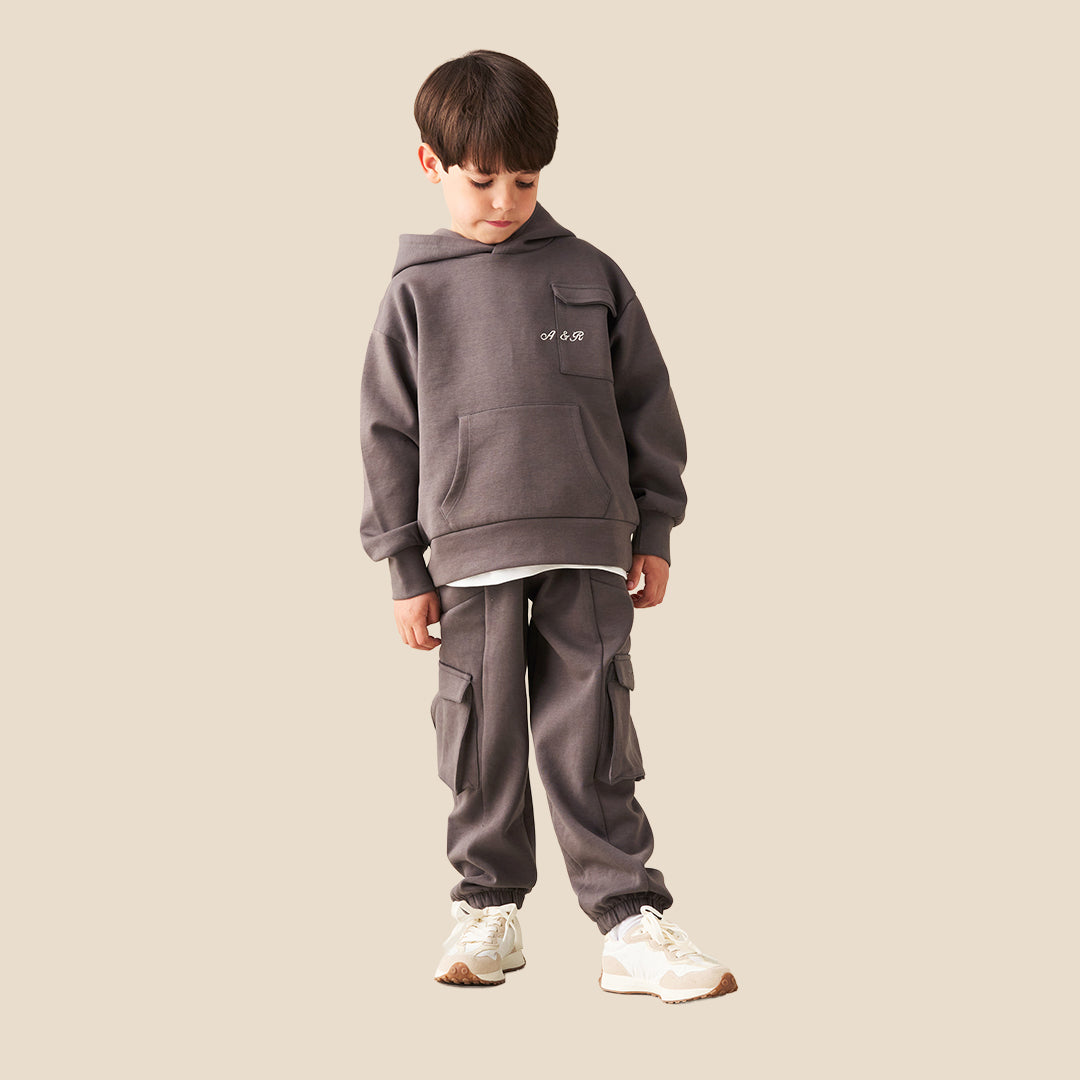 A young boy wearing a dark gray hoodie with a small white graphic on the chest and matching dark gray cargo pants, standing against a solid beige background.