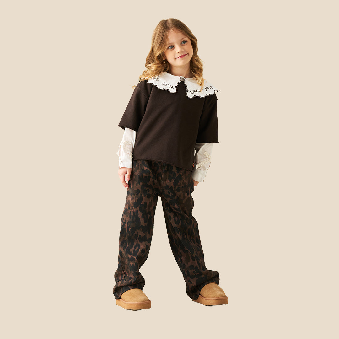 A young girl modeling a costume outfit consisting of a dark brown top with a white embroidered Peter Pan collar, a white long-sleeve shirt underneath, dark brown leopard-print pants, and tan slip-on shoes, set against a solid beige background.