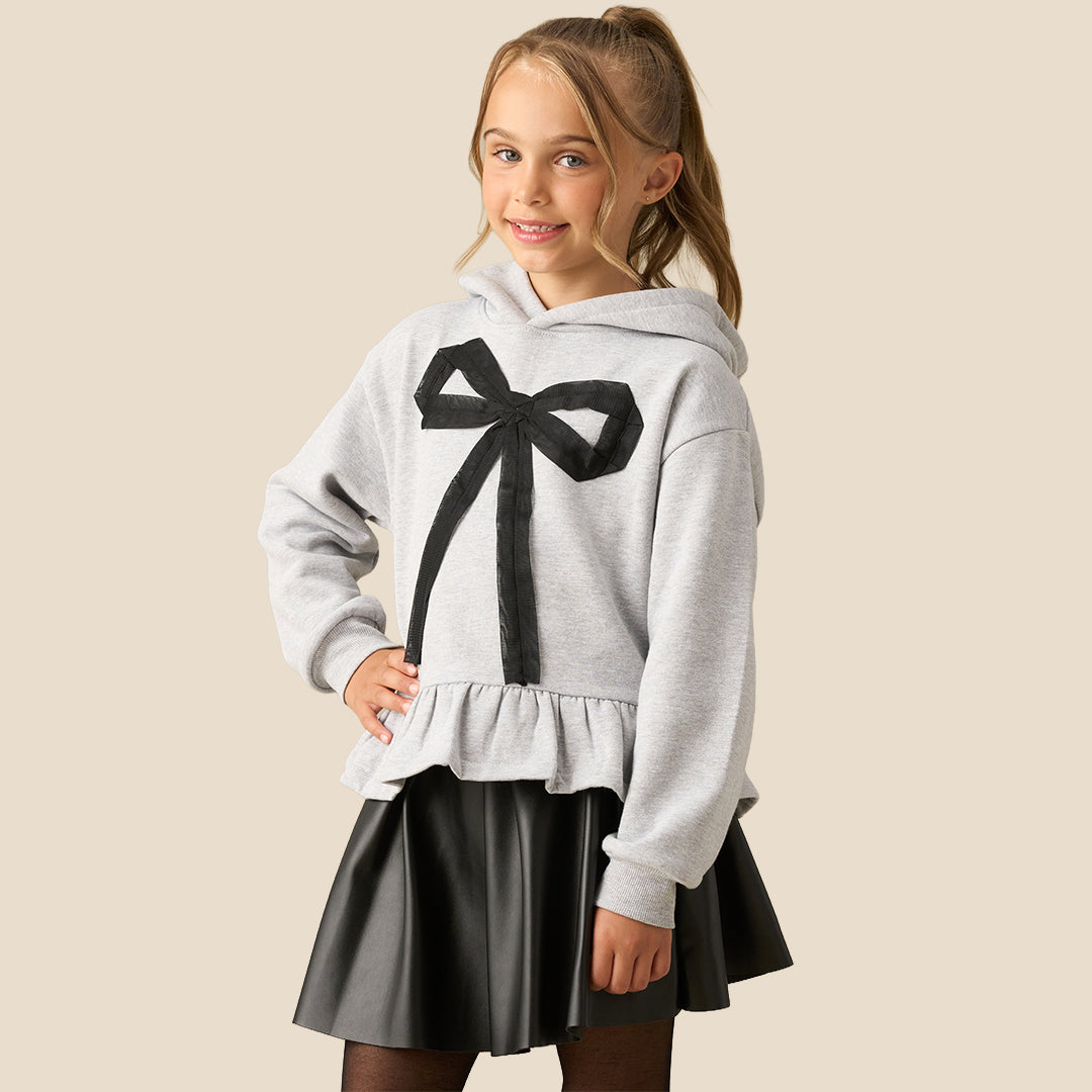 A young girl with her hair in a ponytail models a light gray heathered hoodie featuring a large black bow graphic on the front and a ruffled hem, paired with a black faux-leather skirt, against a solid beige background.
