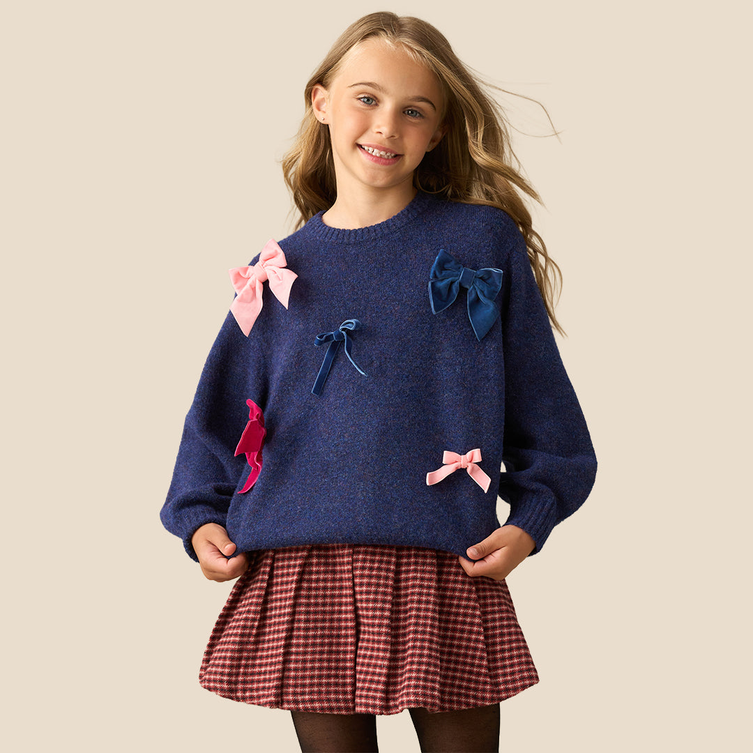 A young girl with blonde hair is smiling and posing in a navy blue knit sweater adorned with pink and blue fabric bows, paired with a red and black gingham pleated skirt, set against a solid beige background.
