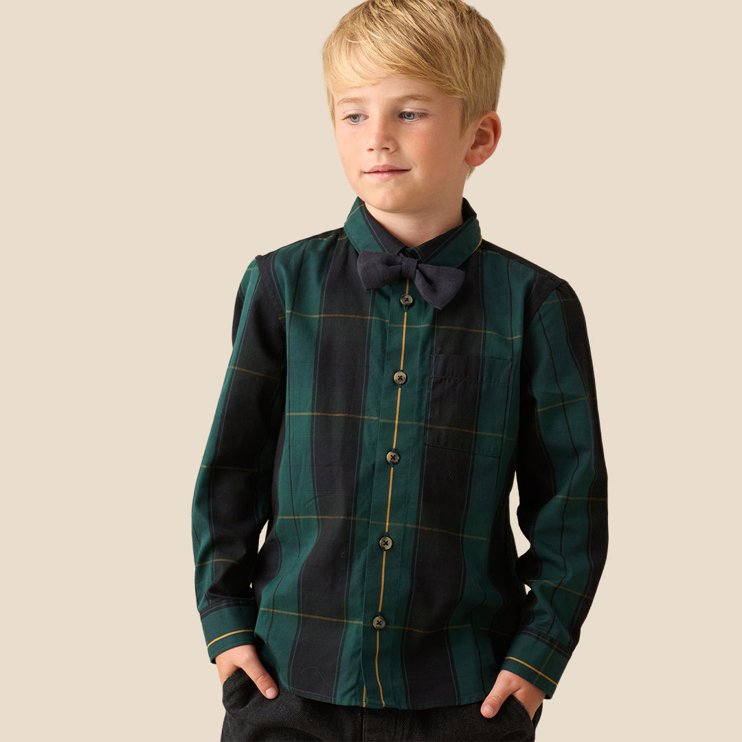 A young blond boy wearing a long-sleeve button-up shirt with a dark green and black plaid pattern, a matching dark bow tie, and dark pants, standing against a solid beige background.