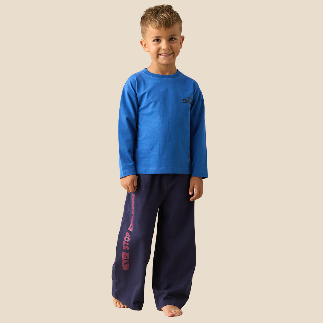 A young boy modeling a two-piece pajama set consisting of a bright blue long-sleeve shirt and navy blue wide-leg pants with pink text on the leg, set against a solid beige background.