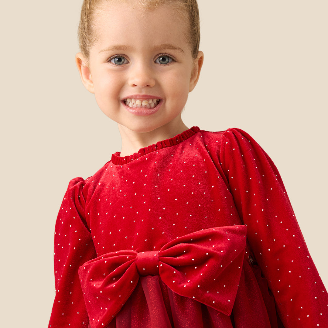 A young girl with her hair in a bun stands against a solid beige background wearing a festive red dress with a polka-dot velvet bodice, long sleeves, a large matching bow at the waist, and a tulle skirt. She is also wearing white knit tights and metallic gold shoes.
