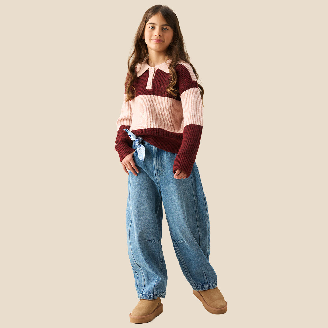 A young girl modeling a ribbed knit polo sweater with horizontal stripes in burgundy and light pink, paired with wide-leg blue jeans against a solid beige background.