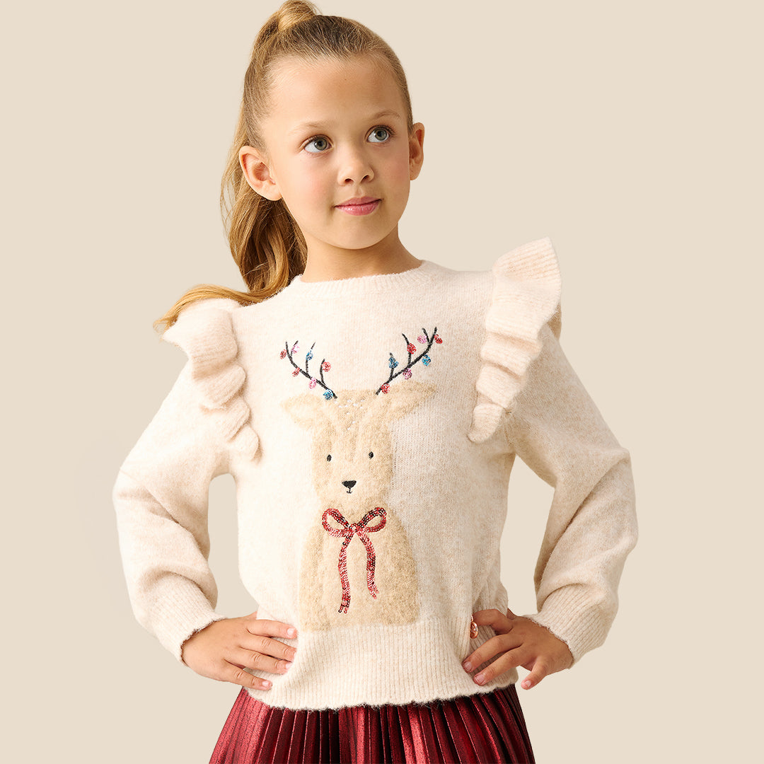 A young girl with blonde hair in a ponytail models a cream-colored knit sweater featuring a large embroidered reindeer on the front with colorful antlers and a red bow. She is wearing a shiny red skirt against a solid light beige background.