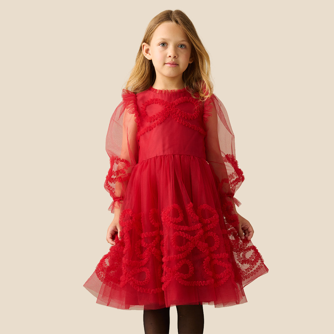 A young girl with blonde hair wearing a red tulle dress with red lace appliques and sheer flutter sleeves, standing against a solid beige background.