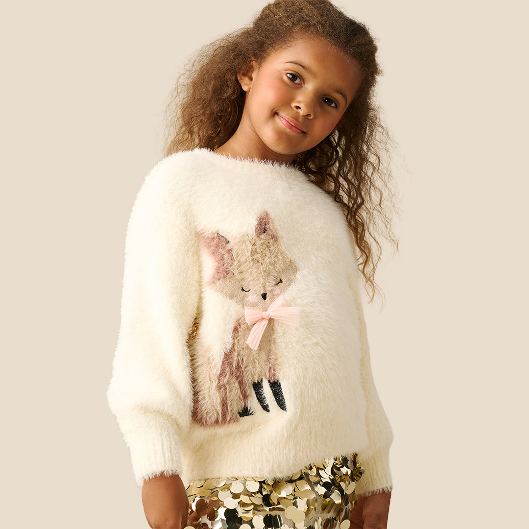 A young girl with curly brown hair wearing a fuzzy off-white knit sweater featuring a sequined fox applique on the front, paired with a gold sequined skirt, set against a solid light beige background.