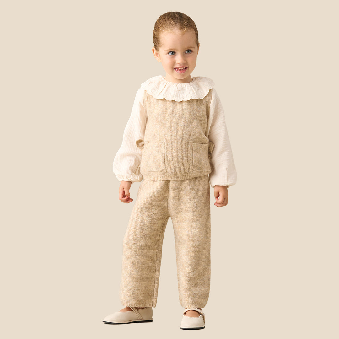 A young child modeling a cream and beige knit outfit, consisting of a long-sleeve top with a ruffled collar and matching knit vest and pants, set against a solid light beige background.