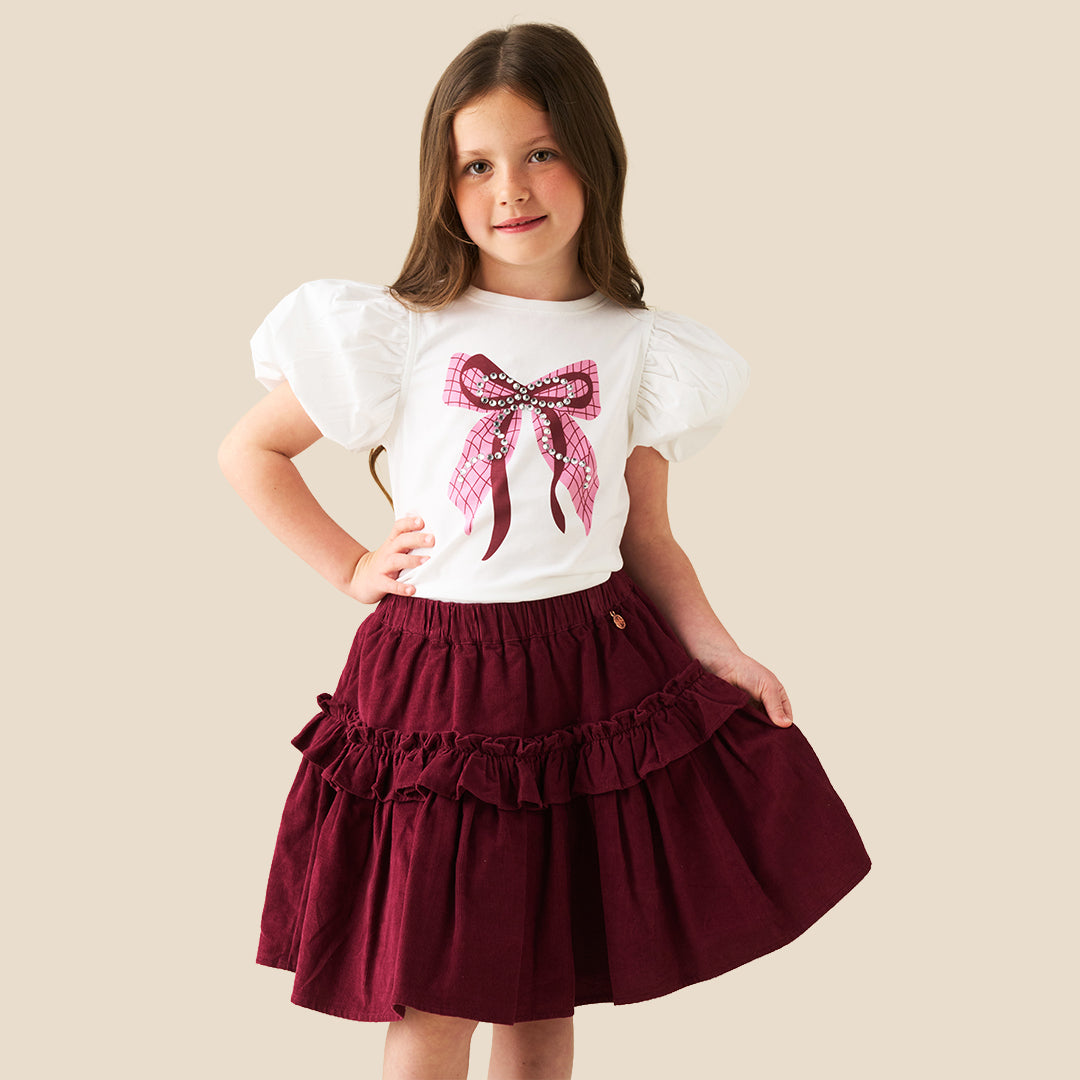 A child wearing a tiered burgundy skirt with ruffles and a white puff-sleeve top featuring a pink plaid bow graphic, photographed against a plain beige background.